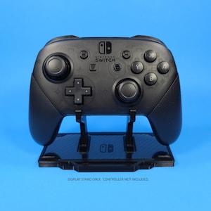 May include: A black Nintendo Switch controller stand with a carbon fiber pattern. The stand is designed to hold a Nintendo Switch Pro Controller. "DISPLAY STAND ONLY. CONTROLLER NOT INCLUDED." is printed below the stand.