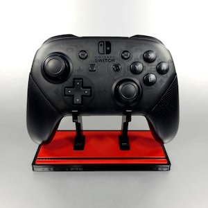 May include: A black Nintendo Switch controller with a red and black stand. The controller has a textured surface and the words "Nintendo Switch" are printed on the front.