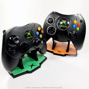 May include: Two black Xbox controllers on display stands. The stands are green and orange. The controllers have the Xbox logo on them.