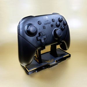 Puede incluir: Un mando negro de Nintendo Switch con un soporte dorado con un diseño de Trifuerza. El mando tiene el logotipo de Nintendo Switch y botones etiquetados como A, B, X, Y y L.