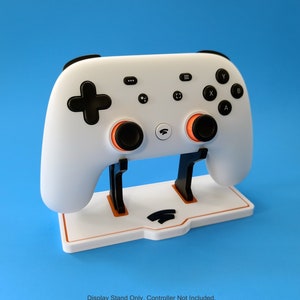 May include: White Google Stadia controller on a white display stand. The stand has a raised Google Stadia logo. "Display Stand Only. Controller Not Included." is printed below the stand.