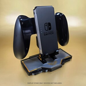 May include: A black Nintendo Switch display stand with attached Joy-Con grips. The stand features a gray and gold base with a Zelda-themed design. The Nintendo Switch console is visible in the center. The text "DISPLAY STAND ONLY. DEVICES NOT INCLUDED." is at the bottom.