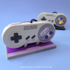 May include: A purple display stand for a Super Nintendo Entertainment System controller. The stand is made of plastic and has a gray base with a purple top. The stand is designed to hold the controller in a vertical position. The text "SUPER NINTENDO ENTERTAINMENT SYSTEM" is printed on the controller.