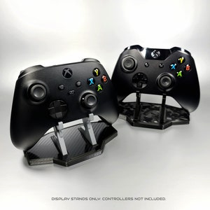 May include: Two black video game controllers on display stands. The stands are made of a black and gray material with a carbon fiber pattern. The controllers are both facing forward with the buttons visible.