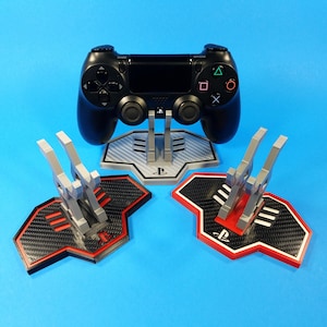 Puede incluir: Tres soportes para mandos de PlayStation impresos en 3D. Dos soportes son negros con detalles rojos y un soporte es gris con detalles blancos. Los soportes están diseñados para mantener un mando de PlayStation en posición vertical.