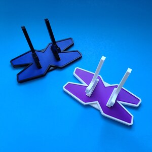 May include: Two 3D printed display stands in the shape of an X. One stand is purple and the other is blue. Both stands have three prongs for holding objects.