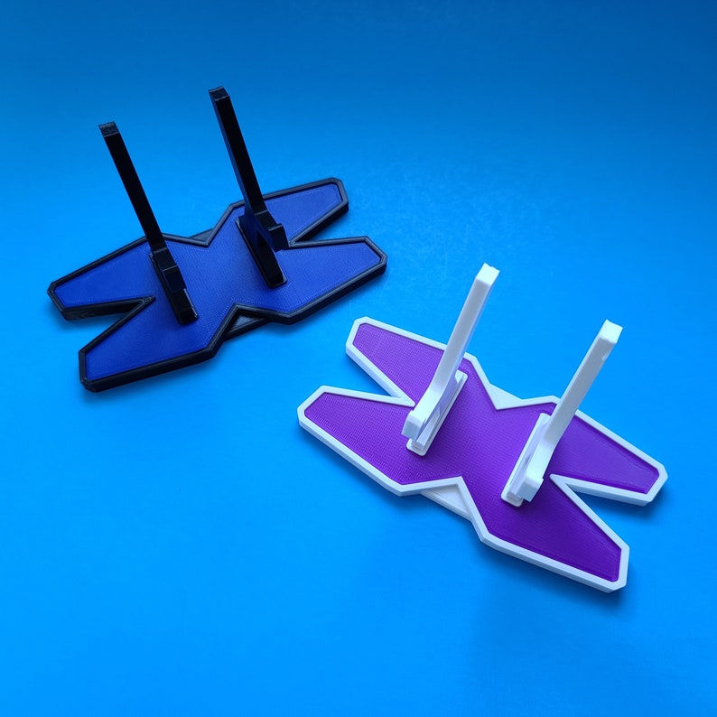 May include: Two 3D printed display stands in the shape of an X. One stand is purple and the other is blue. Both stands have three prongs for holding objects.