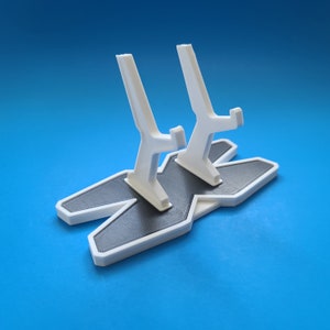 May include: White plastic stand with two slots for holding controllers. The stand is shaped like an X and has a black base.