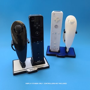 Pode incluir: Três suportes de plástico preto para exibir controles remotos Nintendo Wii. Os suportes são etiquetados com "Wii" e possuem uma ranhura para um controle remoto Wii e uma ranhura para um Nunchuk Wii. Os suportes estão sobre uma superfície azul.