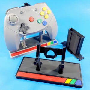 May include: A black and gray 3D printed stand for a Nintendo 64 controller. The stand has two vertical supports and a horizontal bar. The stand is on a gray base with a red stripe and a multi-colored stripe.