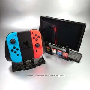 May include: A black stand with a carbon fiber pattern holds a Nintendo Switch console and a Joy-Con controller. The stand also has slots for game cartridges. The console is displaying a black screen.