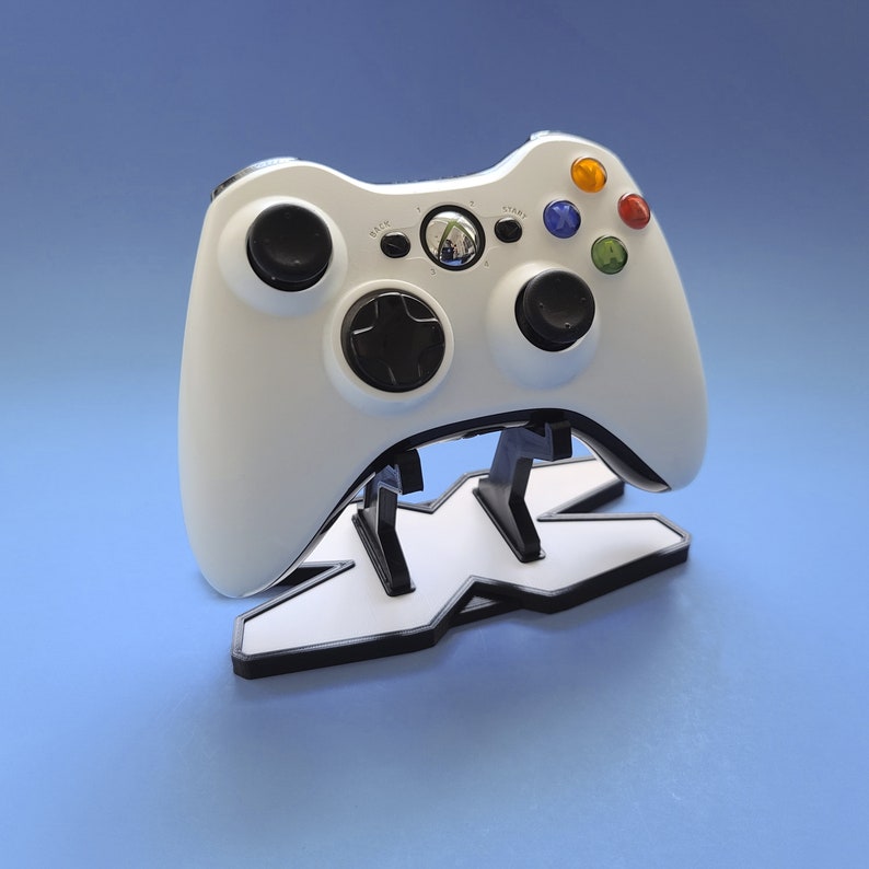 May include: A white Xbox controller with black buttons and a white stand with a black "X" logo. The controller has a black d-pad and black analog sticks.