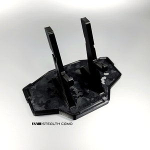 May include: Black plastic phone stand with two slots for phones. The stand has a camouflage pattern and the text "STEALTH CAMO" printed on the bottom.