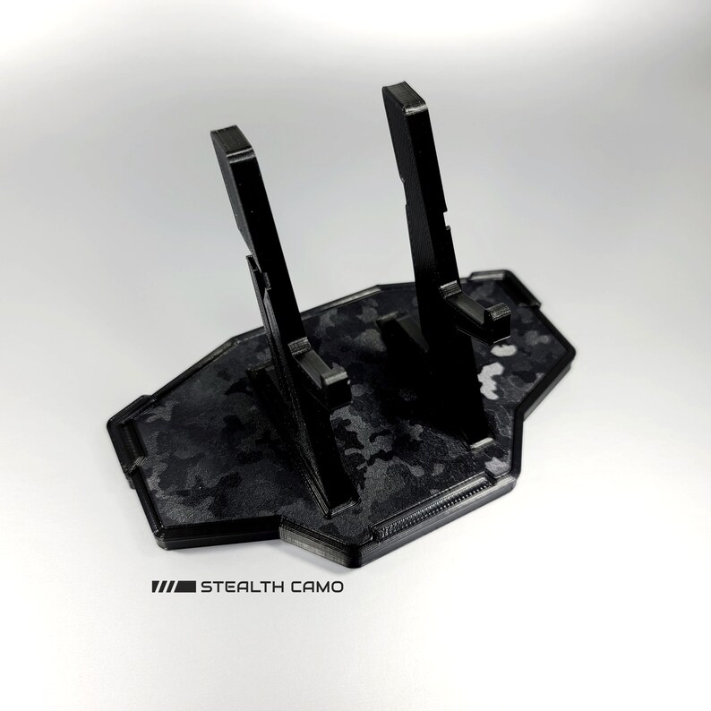 May include: Black plastic phone stand with two slots for phones. The stand has a camouflage pattern and the text "STEALTH CAMO" printed on the bottom.