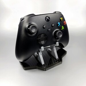 May include: A black Xbox controller on a black and grey carbon fibre stand. The controller has coloured buttons labelled X, Y, A, and B.