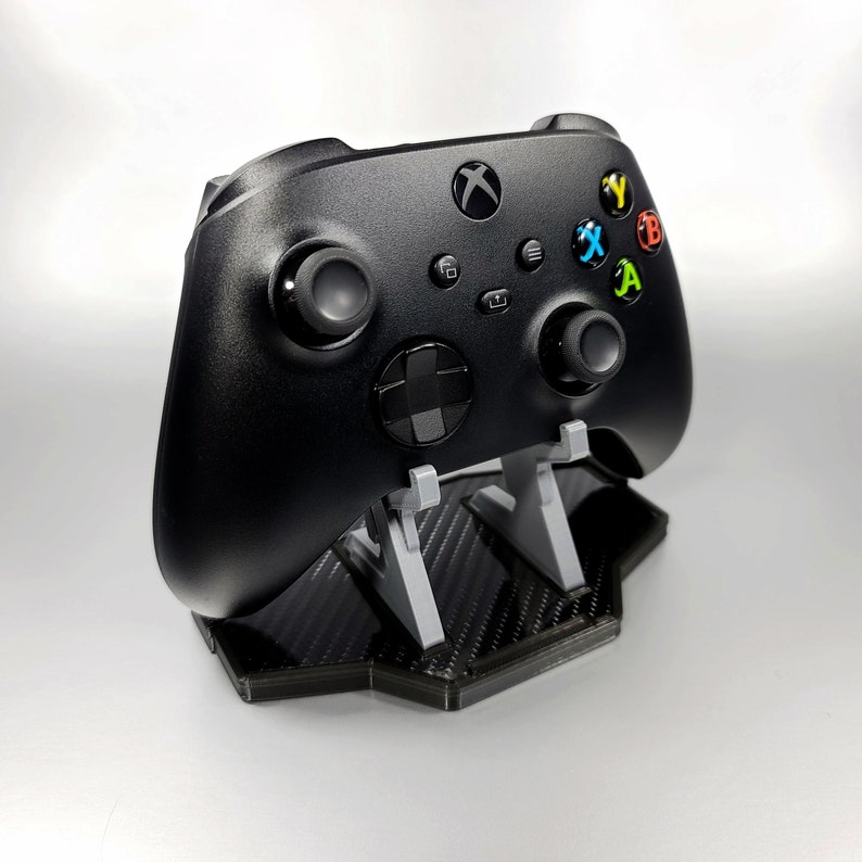 May include: A black Xbox controller on a black and grey carbon fibre stand. The controller has coloured buttons labelled X, Y, A, and B.
