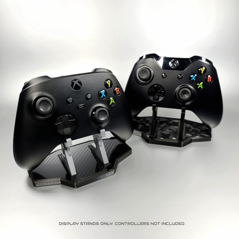 May include: Two black video game controllers on display stands. The stands are made of a black and grey material with a carbon fibre pattern. The controllers are both facing forward with the buttons visible.