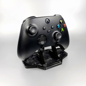 May include: A black Xbox controller with a camouflage-patterned charging stand. The controller has a black and white button layout with green and yellow accents.