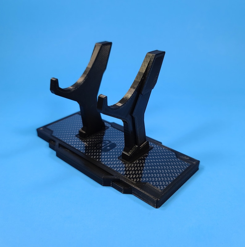 May include: A black plastic stand with a carbon fiber pattern for holding two controllers. The stand has two raised arms with a curved shape.
