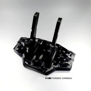 May include: Black 3D printed stand with a carbon fibre pattern. The stand has two vertical supports and a base. The text "FORGED CARBON" is printed on the base.