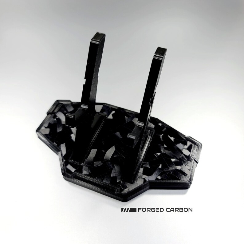 May include: Black 3D printed stand with a carbon fibre pattern. The stand has two vertical supports and a base. The text "FORGED CARBON" is printed on the base.