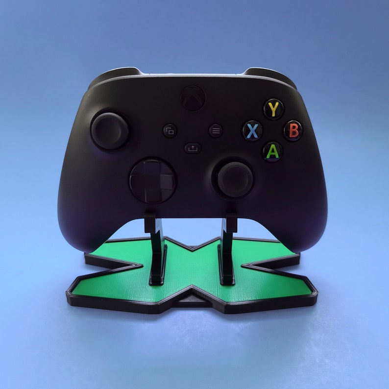 May include: A black Xbox controller with green and black X logo stand. The controller has buttons labeled A, B, X, and Y.