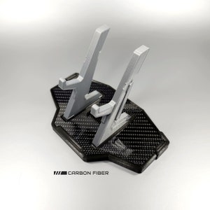 May include: A grey 3D printed phone stand with two slots for phones. The stand has a black base with a carbon fibre pattern and the text "CARBON FIBER" printed on the base.