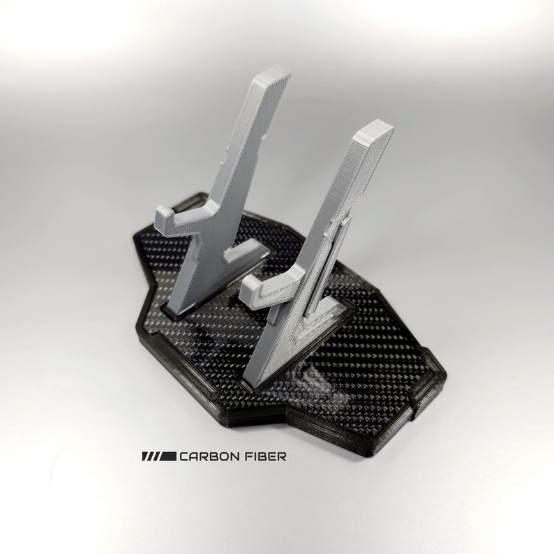May include: A grey 3D printed phone stand with two slots for phones. The stand has a black base with a carbon fibre pattern and the text "CARBON FIBER" printed on the base.