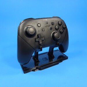 May include: A black Nintendo Switch controller is displayed on a black stand with a carbon fiber pattern. The controller has the Nintendo Switch logo and buttons labeled A, B, X, Y.