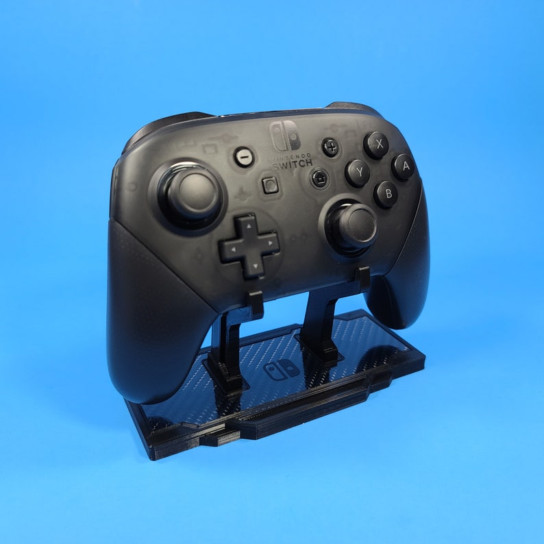 May include: A black Nintendo Switch controller is displayed on a black stand with a carbon fiber pattern. The controller has the Nintendo Switch logo and buttons labeled A, B, X, Y.