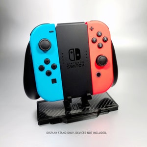 May include: A black, blue, and red Nintendo Switch console display stand with a carbon fiber pattern. "DISPLAY STAND ONLY. DEVICES NOT INCLUDED." is printed below the stand.