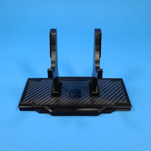 May include: Black plastic stand with a carbon fiber pattern and two raised arms for holding video game controllers. The stand has the Nintendo Switch logo in the center.