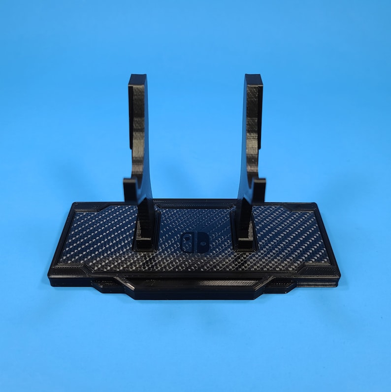 May include: Black plastic stand with a carbon fiber pattern and two raised arms for holding video game controllers. The stand has the Nintendo Switch logo in the center.