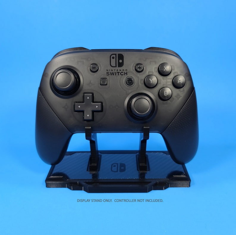 May include: A black Nintendo Switch controller stand with a carbon fiber pattern. The stand is designed to hold a Nintendo Switch Pro Controller. "DISPLAY STAND ONLY. CONTROLLER NOT INCLUDED." is printed below the stand.