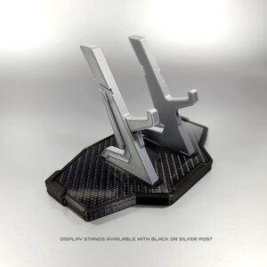 May include: A 3D printed display stand with a black carbon fibre base and two silver posts. The stand is designed to hold two items, such as video game controllers or figurines. Display stands available with black or silver post.
