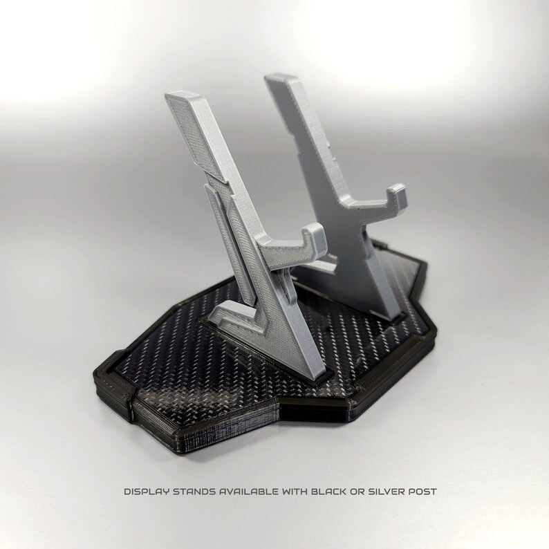 May include: A 3D printed display stand with a black carbon fibre base and two silver posts. The stand is designed to hold two items, such as video game controllers or figurines. Display stands available with black or silver post.