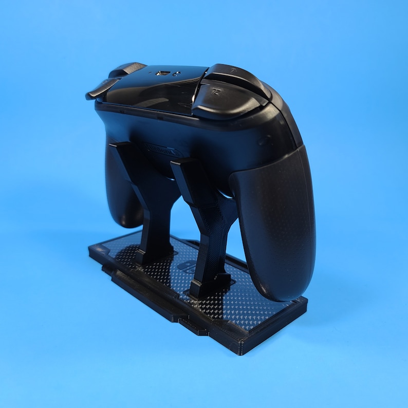 May include: A black video game controller stand with a carbon fiber pattern. The stand is designed to hold a controller upright.