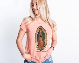 May include: A peach-colored V-neck t-shirt featuring an image of Our Lady of Guadalupe. The image is framed by a gold-colored oval and depicts a religious figure in a teal robe. The text "Our Lady of Guadalupe" is printed below the image.