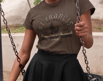 May include: Brown t-shirt with a round neck featuring a graphic of a wolf and the text "ST. FRANCIS OF ASSISI, Patron Saint of Animals, Wolf Tamer of Gubbio, ITALY". The shirt is paired with a black skirt.