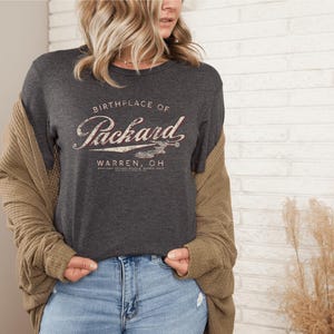 May include: Dark gray t-shirt with the text "Birthplace of Packard Warren, OH" in a vintage style. The design includes an airplane graphic. The shirt is paired with blue jeans and a tan cardigan.