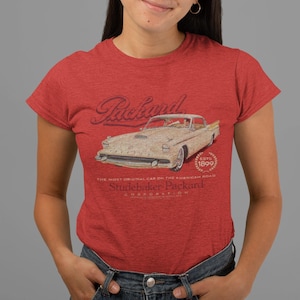 May include: Red t-shirt featuring a vintage Packard car graphic. The design includes the Packard logo, the text "ESTO 1899", and the phrase "THE MOST ORIGINAL CAR ON THE AMERICAN ROAD". The shirt is made of a soft material.