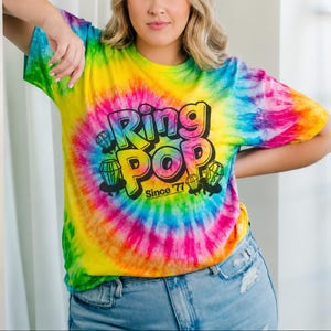 May include: A tie-dye t-shirt with a vibrant swirl of rainbow colors. The shirt features the text "Ring Pop" in a bold, outlined font, with diamond graphics and the text "Since '77". The shirt is a short-sleeved crew neck.