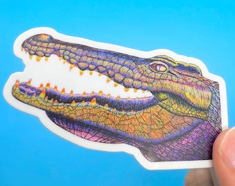 Pode incluir: Um adesivo colorido de um jacaré com a boca aberta, mostrando dentes afiados. O jacaré é desenhado em um estilo detalhado com um esquema de cores roxo, azul e amarelo.