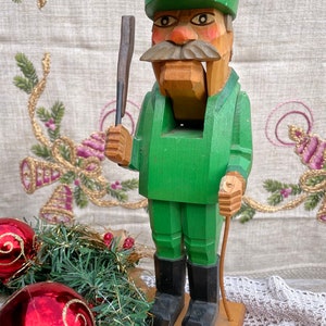 May include: A wooden nutcracker figurine of a man in a green outfit, hat, and black boots, holding a tool and a walking stick. The figurine is on a wooden base. Christmas ornaments and garland are in the foreground.
