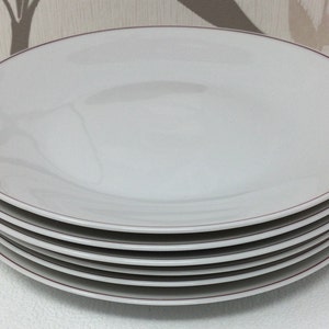 May include: A stack of six white dinner plates with a thin red rim.