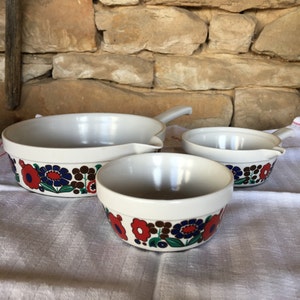 May include: Three white ceramic bowls with a floral design in red, blue, green, and brown. The bowls have a pouring spout on the side.