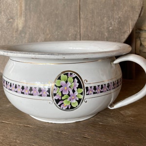May include: White ceramic chamber pot with a floral design featuring purple flowers and gold accents. The pot has a handle on the side.