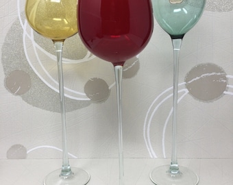 May include: Three tall glass vases, one red, one yellow, and one green. The vases are all clear glass with a long, thin stem and a wide, round base.