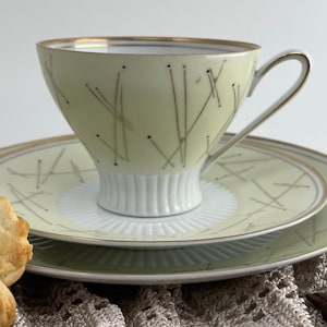Könnte beinhalten: Ein Vintage-Teetassen- und Untertassen-Set mit einem abstrakten Design in Hellgrün und Gold. Das Set befindet sich auf einer gehäkelten Oberfläche mit Keksen und Kaffeebohnen.