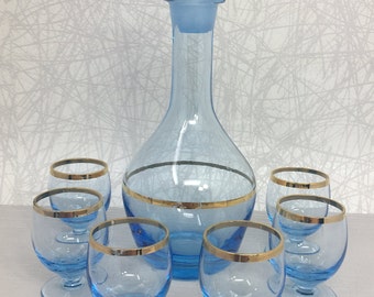 May include: A set of six blue glass liquor glasses with gold trim and a matching blue glass decanter with a gold band around the middle.